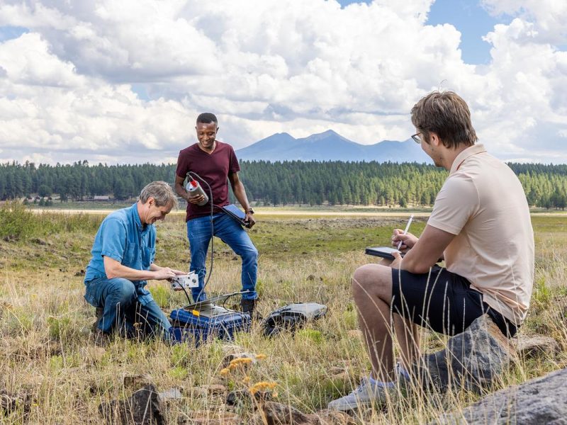 A researcher and two students placing research equipment in the field. The San Francisco Peaks can be seen in the background.