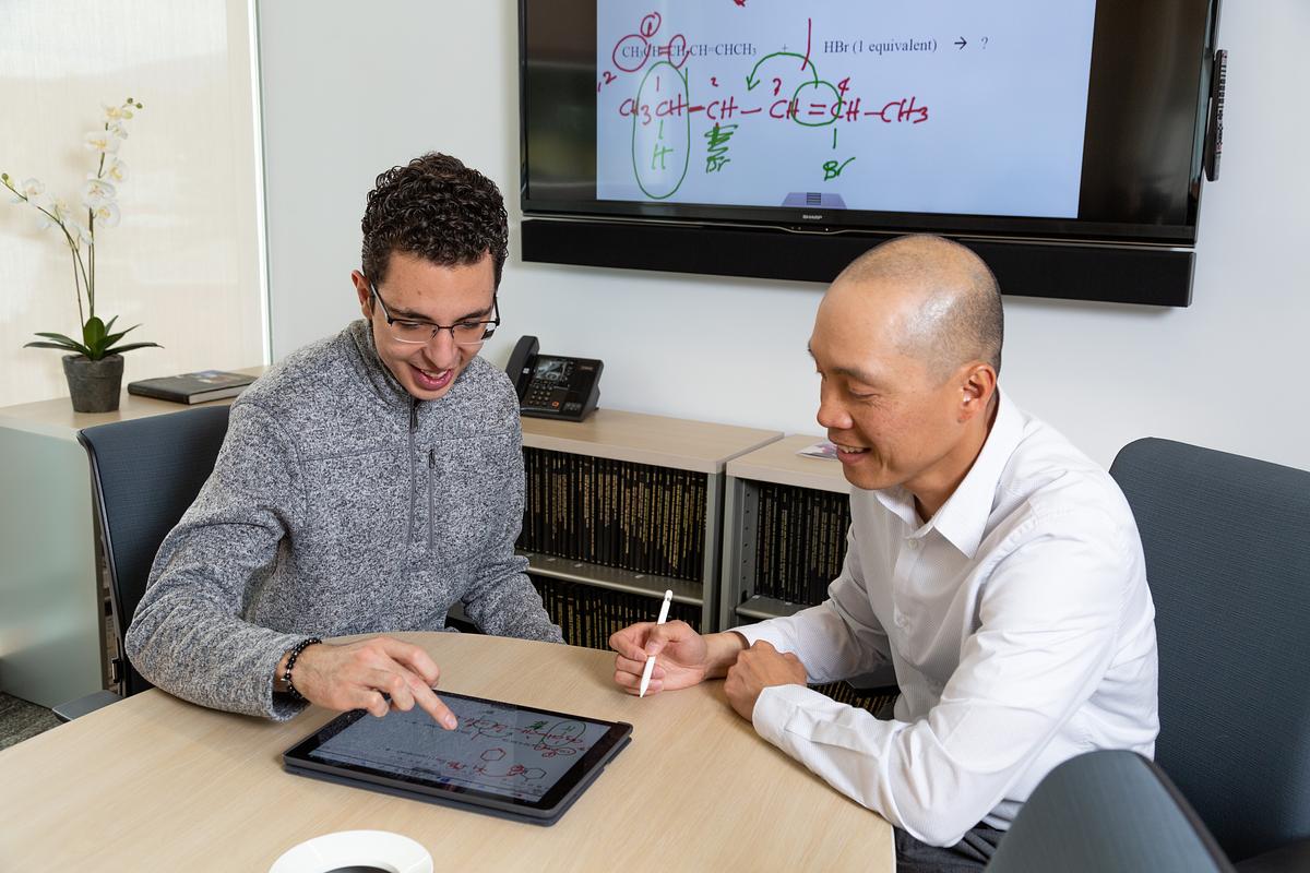 A student and researcher, one wearing a grey sweater and the other wearing a white shirt, are seated at a table in an office setting. A whiteboard with mathematical equations is visible in the background. The two individuals appear to be engaged in a discussion or meeting.