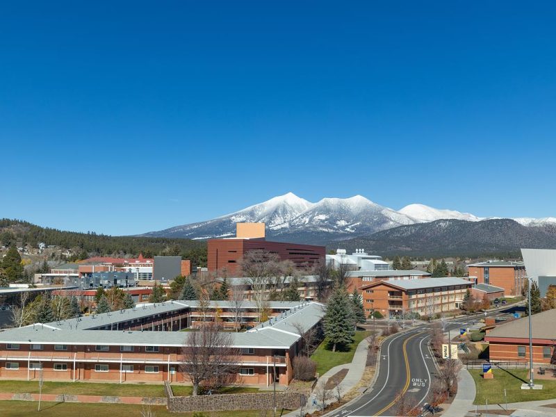 N A U campus buildings on a clear day, snow-capped mountain peak visible in the background, curved roadway and landscaping in foreground, evergreen trees surrounding the area under blue sky.