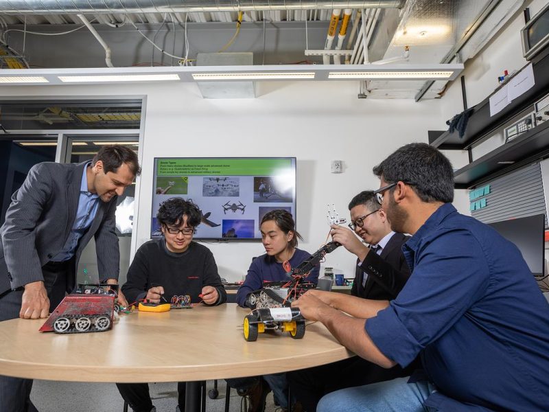 Several students and researcher sitting and some standing, gathered around a table. They appear to be examining and interacting with small model vehicles or robots on the table.