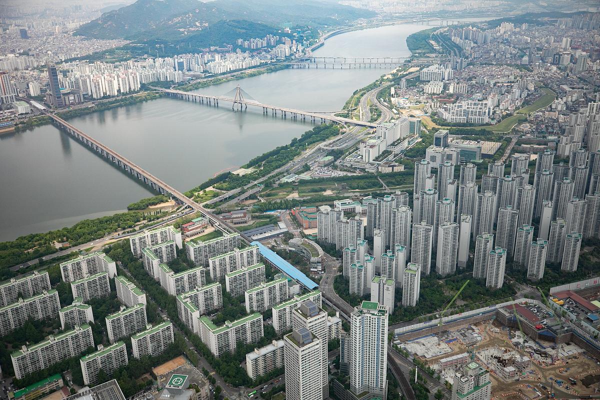 Aerial view of a densely populated urban area with high-rise buildings, bridges, and a river. The cityscape extends over a wide geographical area with mountains visible in the distance.