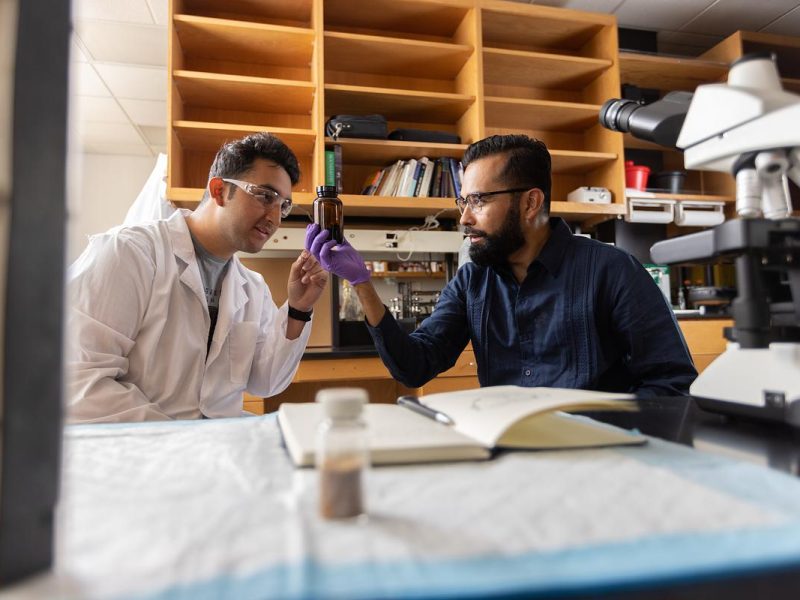 A researcher and student wearing casual clothing, seated at a desk in what appears to be a laboratory or research setting. One person is holding a test tube. Shelves containing books and lab equipment are visible in the background.