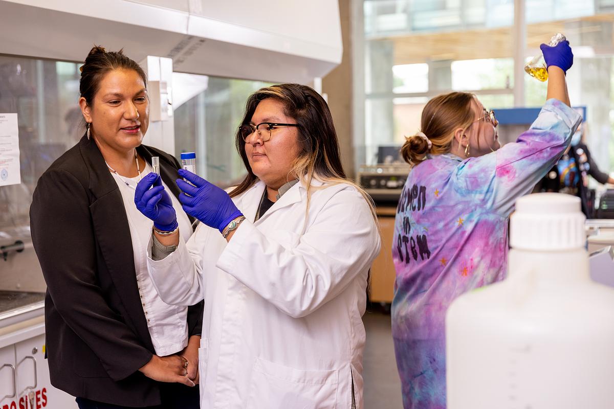 Researchers working with students. Two wearing white lab coats and blue gloves, interacting in a laboratory setting.