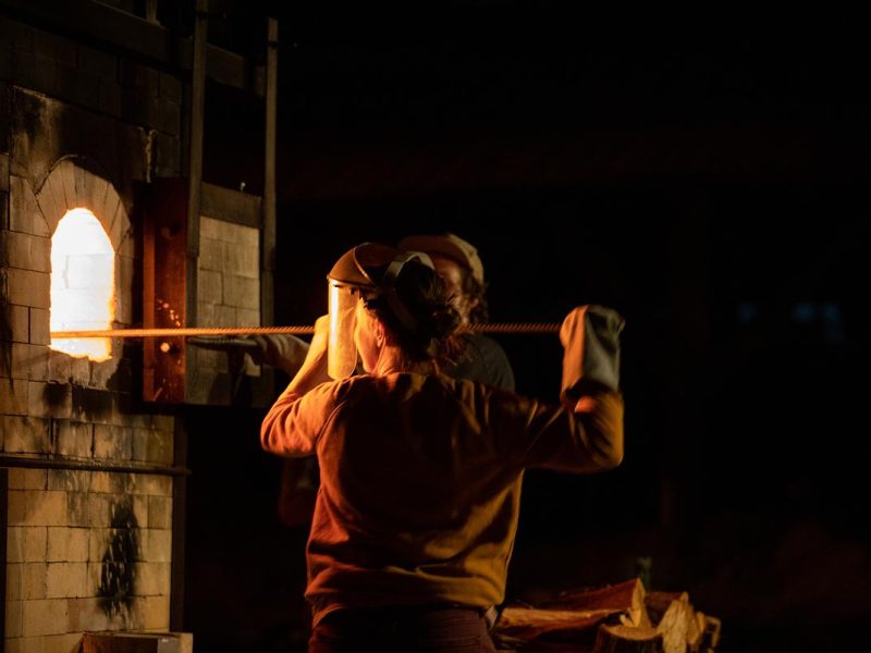 A person in an orange jacket standing near a kiln with the back of their head visible.
