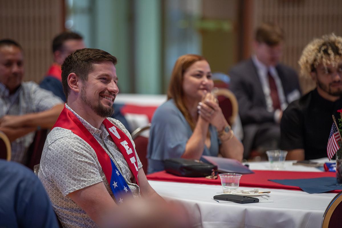 Smiling student sitting at a table wearing a Veteran graduation stole. Other people are sitting around him.