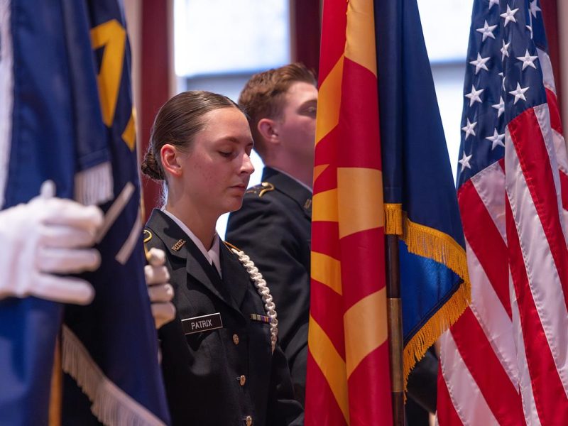 Military students standing in front of flags, including flags of the United States of America and what appears to be a state flag.