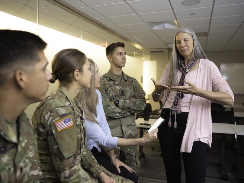 Multiple people in a classroom setting. There are four individuals ROTC students and a researcher standing together talking.