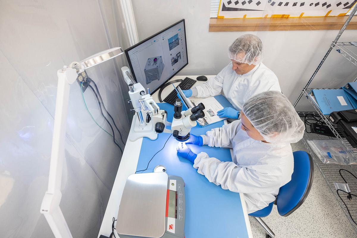 Laura Lee and Dr. Christopher Edwards in protective clothing sitting at a workstation in a research lab with various scientific equipment and instruments, including microscopes and displays showing biological or medical imagery.