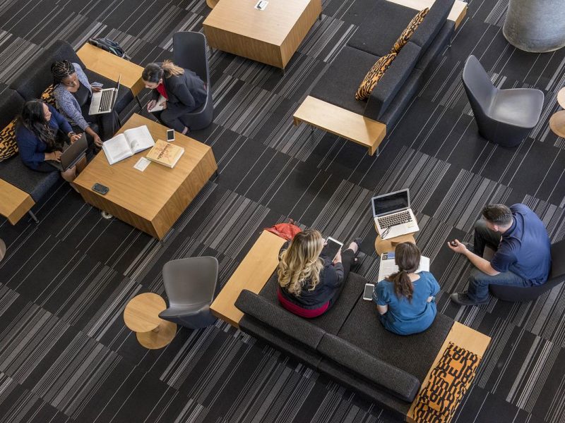 People sitting or standing in a modern office space, using laptops and other digital devices. The setting appears to be a shared workspace or collaborative environment, with various seating arrangements and furnishings. The floor is covered in a striped pattern, and there are some decorative elements visible.