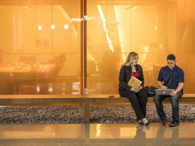 Individuals sitting on a bench. One person is holding a file folder and the other is using a laptop. The setting appears to be an indoor lobby or public space with glass walls and floors.