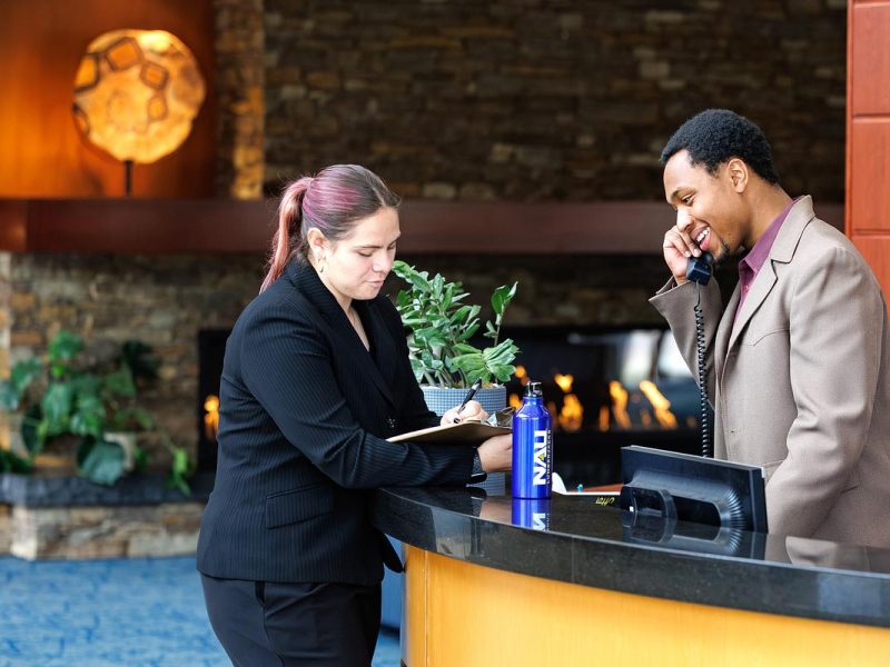 Two people at a hotel reception desk in a modern lobby with stone walls, fireplace, and ambient lighting. The person on the left wears a black blazer and holds a tablet, while the person on the right wears a beige jacket. A potted plant, water bottle, and blue carpet are visible in the professionally designed interior space.