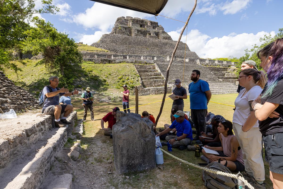 Ruins of a large, ancient stone structure with steps leading up to it, surrounded by lush greenery. Jamie Awe and students sitting and standing around the ruins, examining and exploring the site.