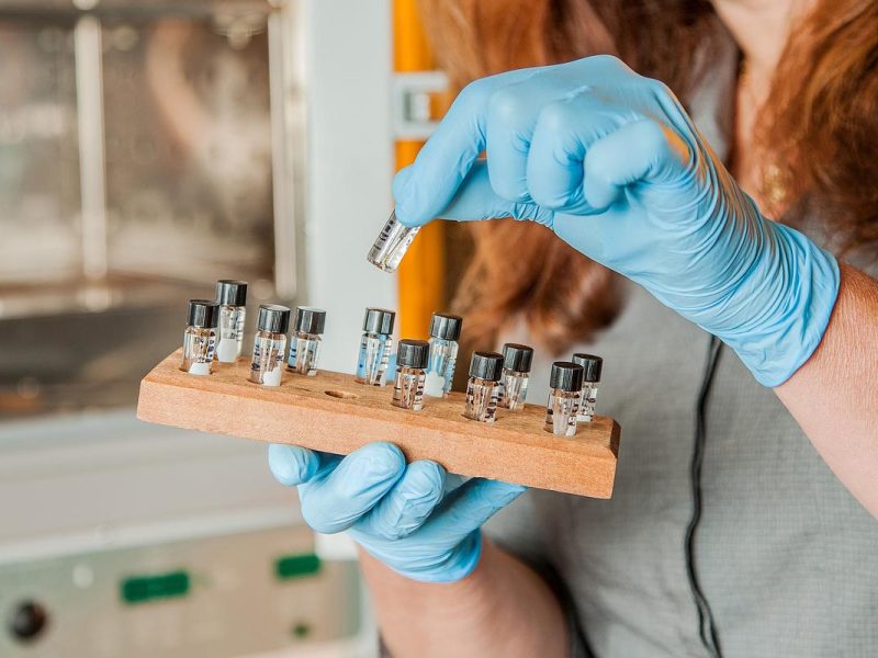 Person wearing blue gloves and placing clear containers with black caps into a wood tray.