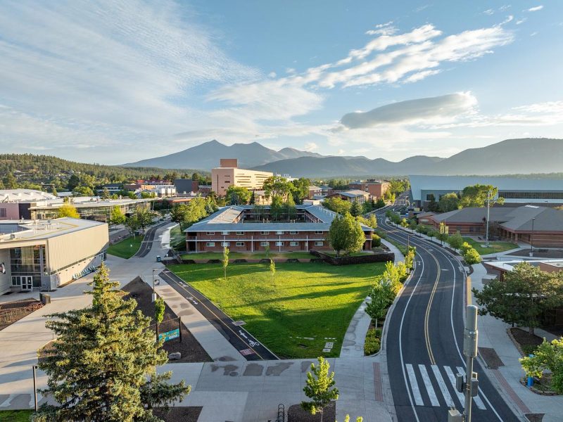 Aerial view of N A U campus with mountains in the background. Multi-story buildings, paved roads, grassy areas, and trees are visible. The sky has clouds and blue color.