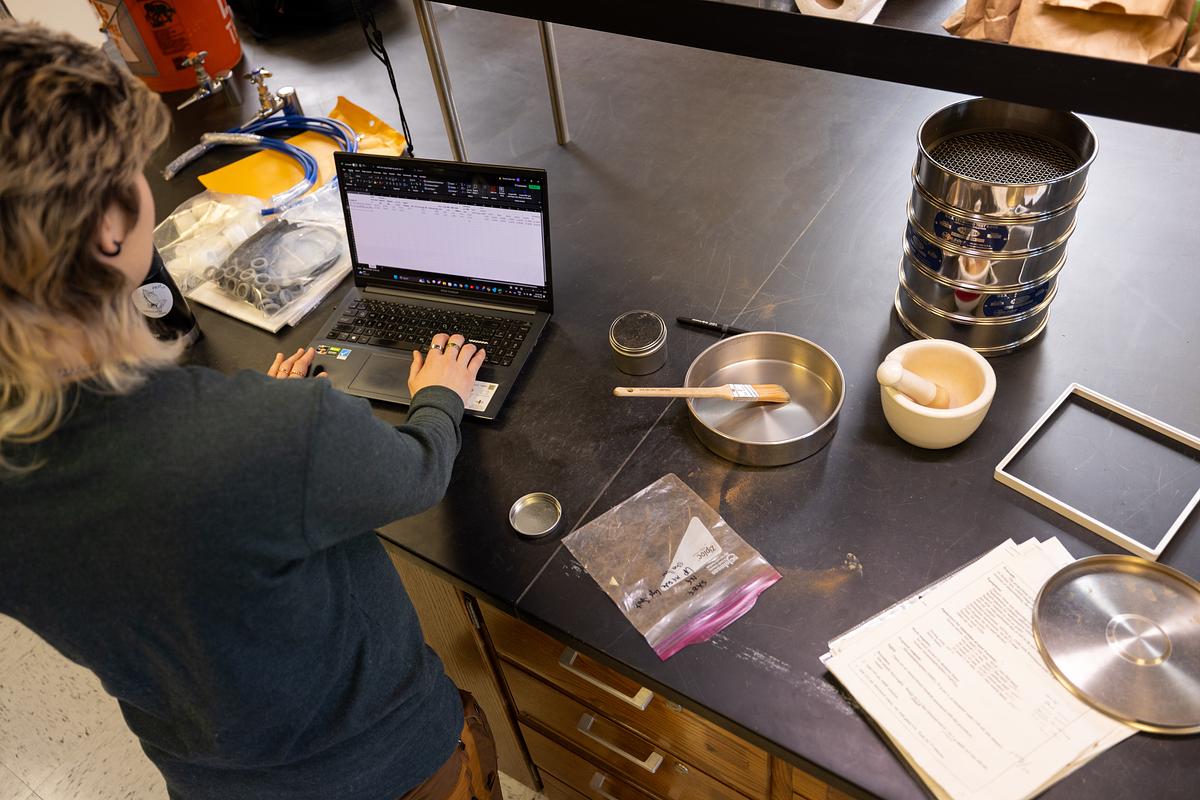 A person sitting at a desk, working on a laptop computer. The desk is cluttered with various items including a tablet device, magnifying glass, and what appears to be drug paraphernalia. There is also a stack of papers and other office supplies visible.