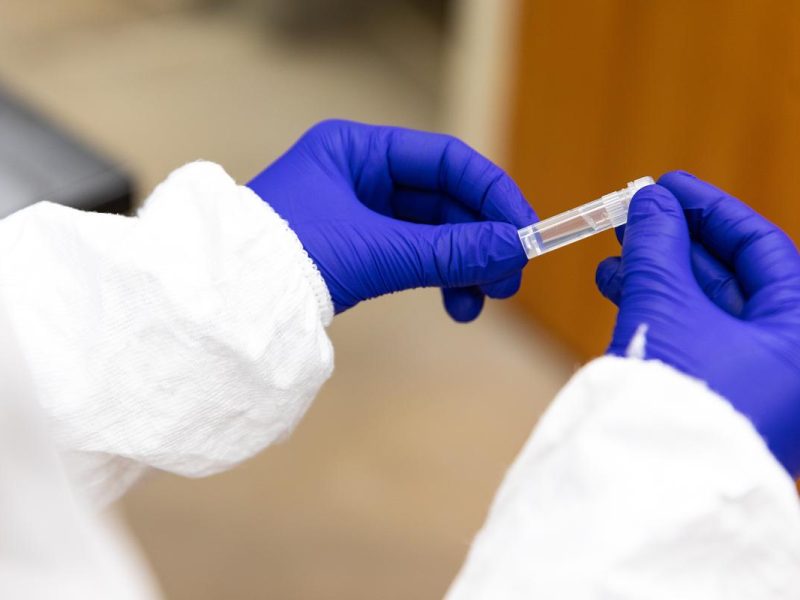 Person wearing a white lab coat and purple gloves holding a vial with liquid.