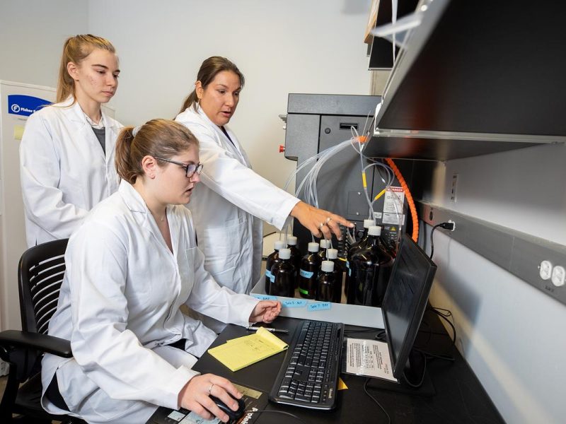 Three individuals in white lab coats working at a computer in a laboratory setting. Shelving and equipment visible in the background.