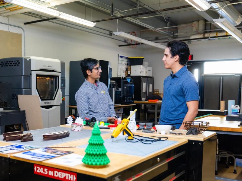 Two people having a conversation in a laboratory workspace with 3D printers and manufacturing equipment. The workbench displays a green 3D printed Christmas tree model with a red label reading "24.5 IN DEPTH" along with various tools and materials. Industrial ceiling with fluorescent lighting and exposed ductwork visible overhead.