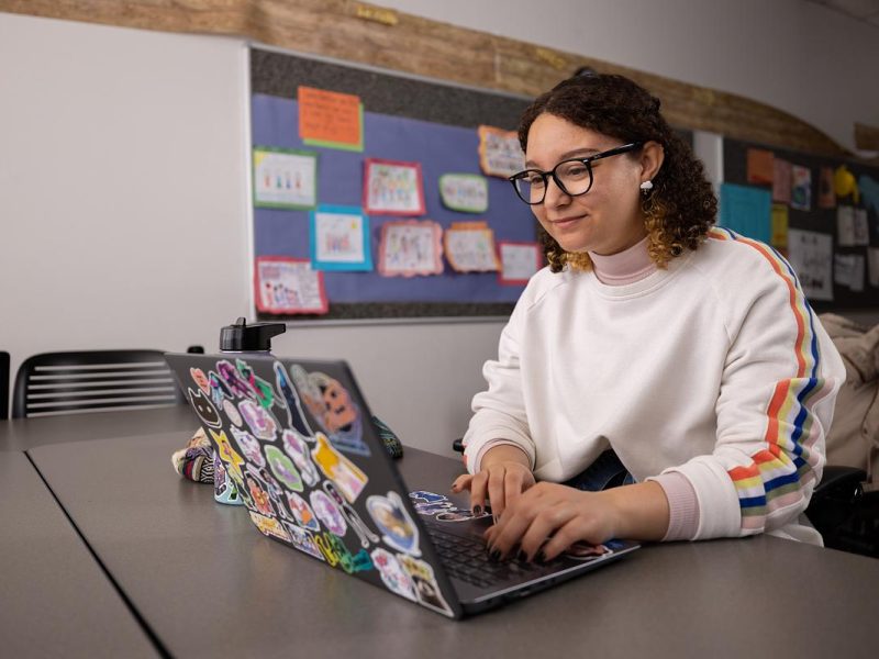 Person sitting at a desk, wearing glasses and a white sweater, typing on a laptop computer. A bulletin board with various papers or notes is visible in the background.