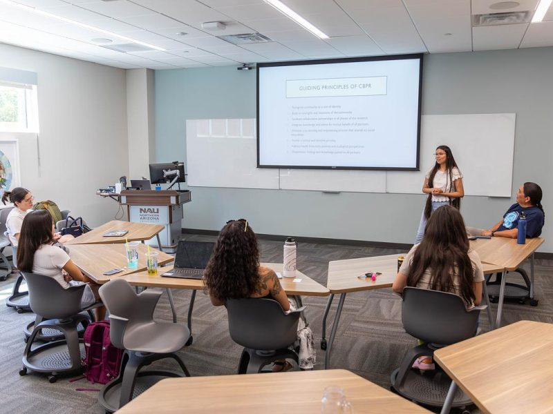 Classroom setting with a presenter standing in front of a group of students seated at tables, with a presentation screen visible.
