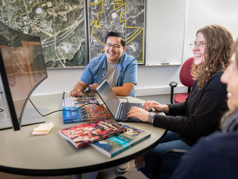 Three people seated around a curved table in an office or classroom setting with maps visible on the wall behind them. A desktop computer monitor is on the left side of the table, and several colorful books or magazines are spread across the table surface. The people appear to be in a collaborative meeting or study session, with at least two individuals working on laptops.