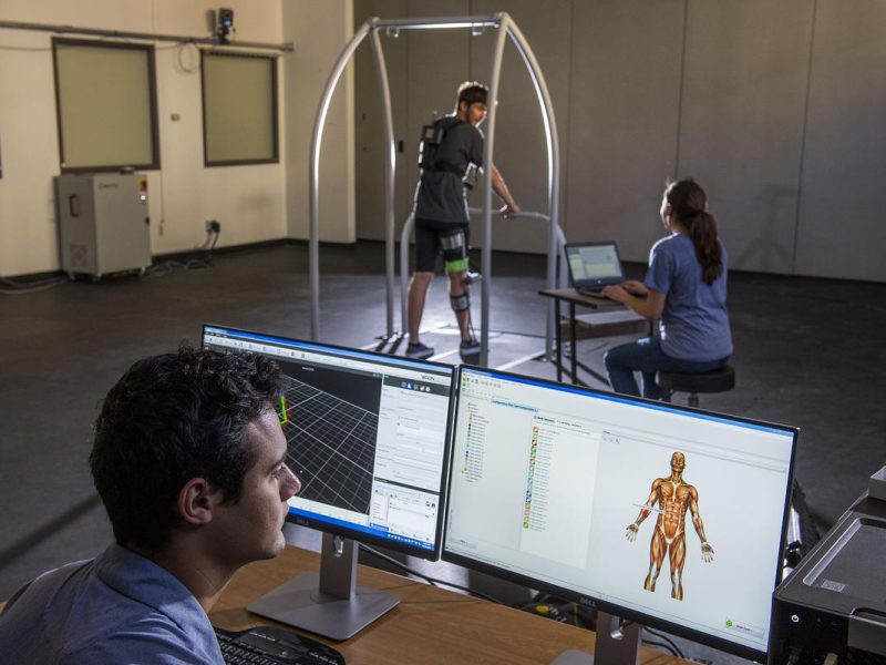 Person sitting in front of two computer screens with a human skeleton displayed. In the background are two people one is on a device to test exoskeleton.