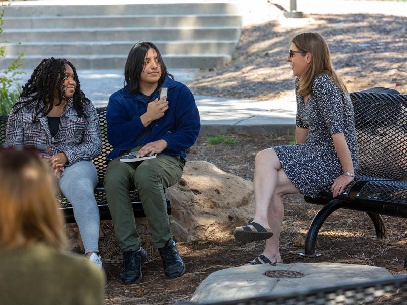 Three individuals, dressed casually, sitting on benches and talking in a natural outdoor setting with trees and rocks visible in the background.