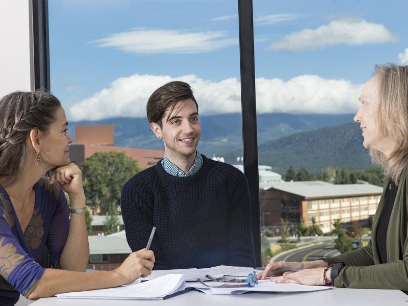 Three people sitting at a table talking with papers and notebooks spread out on the table.