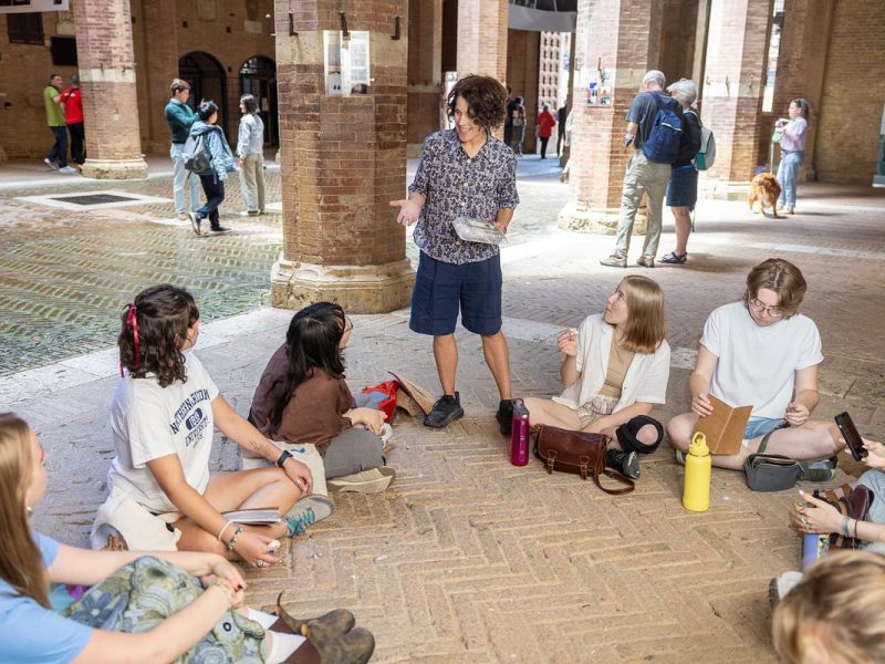 Brick building, people sitting on the ground, outdoor setting, backpacks, water bottles, books, laptops, conversation, interaction.