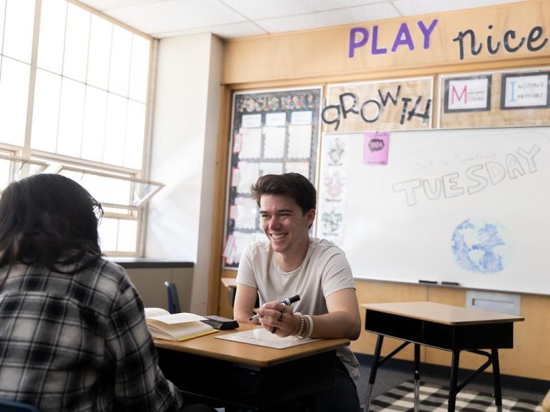 Two people seated at desk in classroom, one smiling and writing, "Play nice, Growth, Tuesday" on whiteboard.