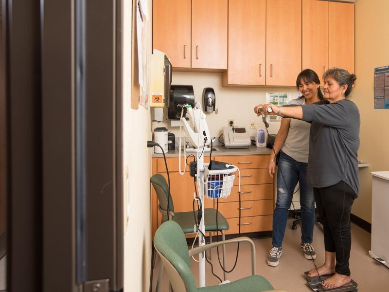 Two people standing in a doctors office. One is standing on a scale and the other is observing her.