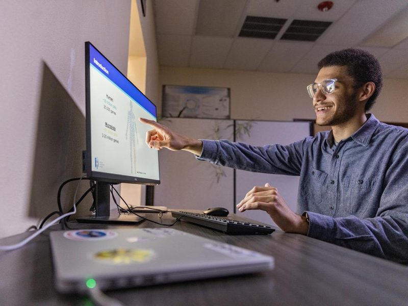 Man in gray shirt sitting at a desk using a computer with a screen displaying text. The man is smiling and using the computer in an office or workplace environment.