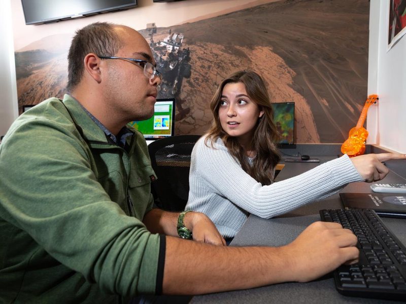 Two people sitting at a desk in chairs. One of them is pointing to something on the computer screen.