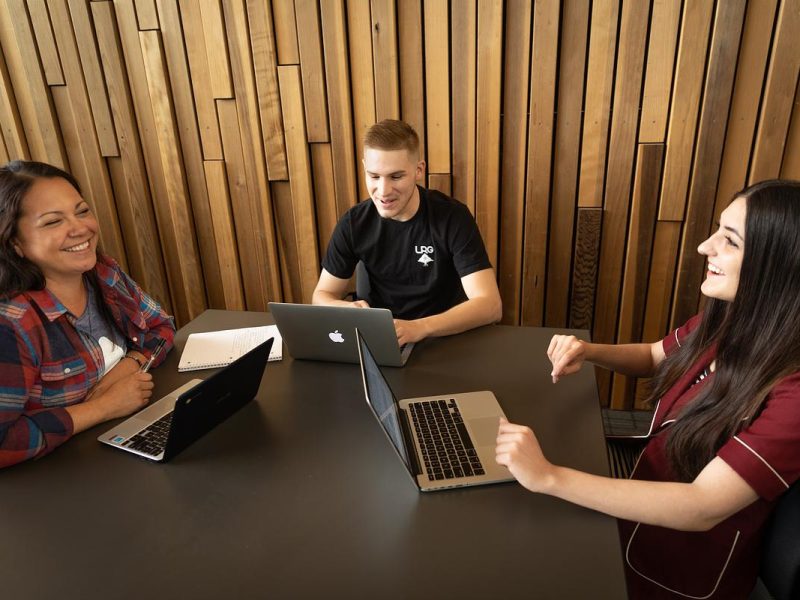Three people sitting around a table, using laptops and appearing to have a discussion. The background is a wooden wall paneling.