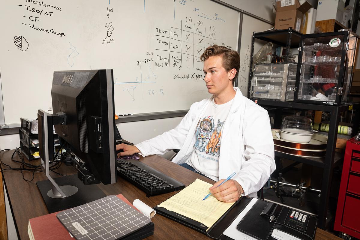 Person wearing a white lab coating sitting at a desk in front of a computer in a lab environment.