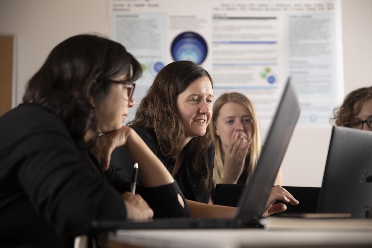 Several individuals seated at desks, using computers. The individuals appear to be focused on their work, with some expressions of concentration or discussion visible. The backdrop includes a presentation board or other informational display. The overall scene suggests an educational or office setting.