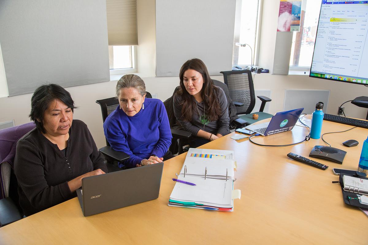 Three researchers sitting at a desk in a conference room. They are working on computers and talking to each other.