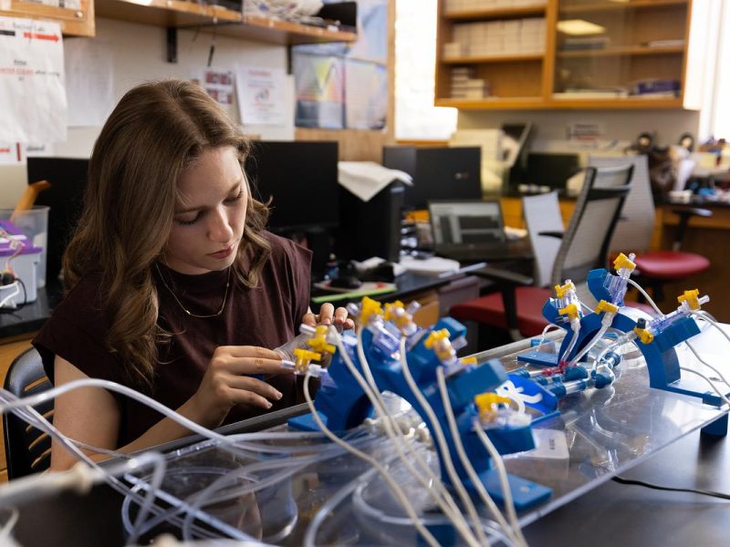 Person sitting at desk examining laboratory equipment, including tubes and devices, surrounded by electronics and computer equipment in an office or workspace setting.