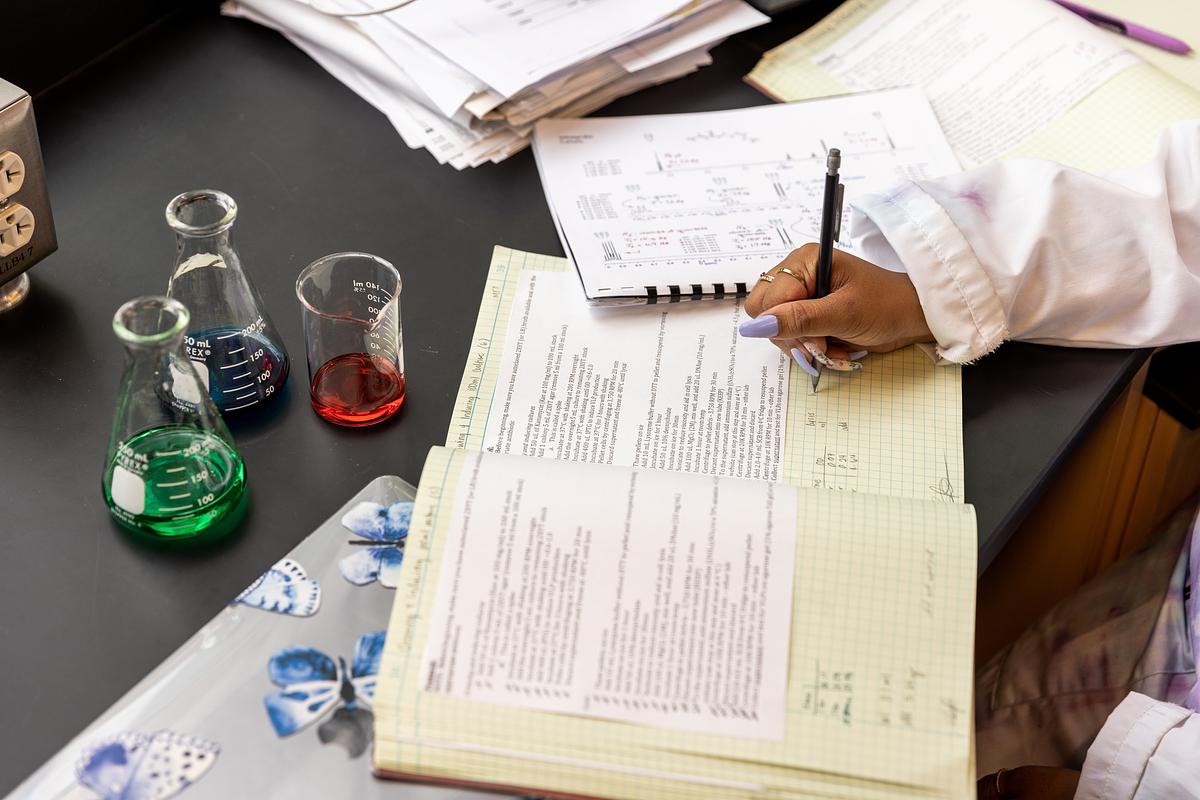 Desk with various scientific equipment, including glass beakers containing colored liquids, and a person's hand writing on a document.