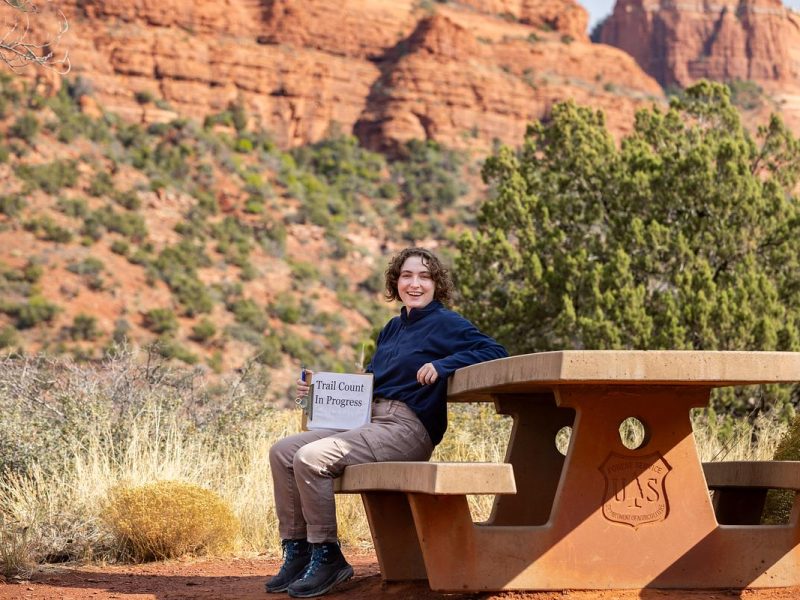 Smiling person sitting on wooden bench in outdoor landscape with red rock formations and green vegetation. "Trail closed in progress" sign visible.