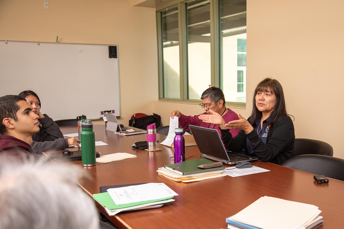 Group of people sitting around a table in an office or classroom setting. They appear to be engaged in a discussion or meeting, using laptops and other materials. The scene depicts a typical workspace environment.