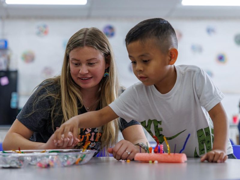 A teacher and child working together at a table in a bright classroom, engaged in a hands-on creative activity with art supplies and markers.