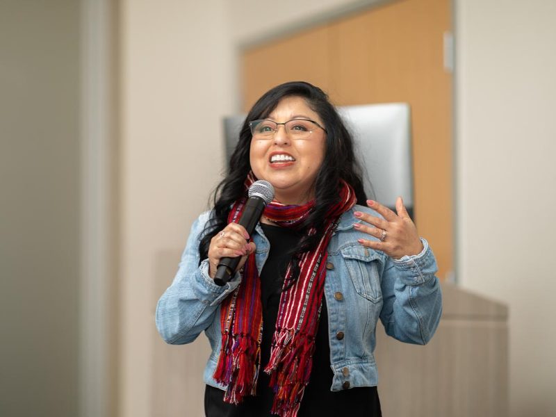 Middle-aged person with long, dark hair wearing a jean jacket, red scarf, and glasses, smiling and speaking into a microphone.