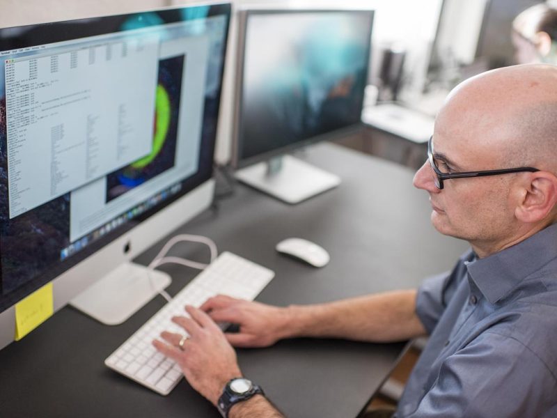 Man wearing glasses, seated at a desk, using a computer keyboard and mouse while looking at a computer screen displaying data.