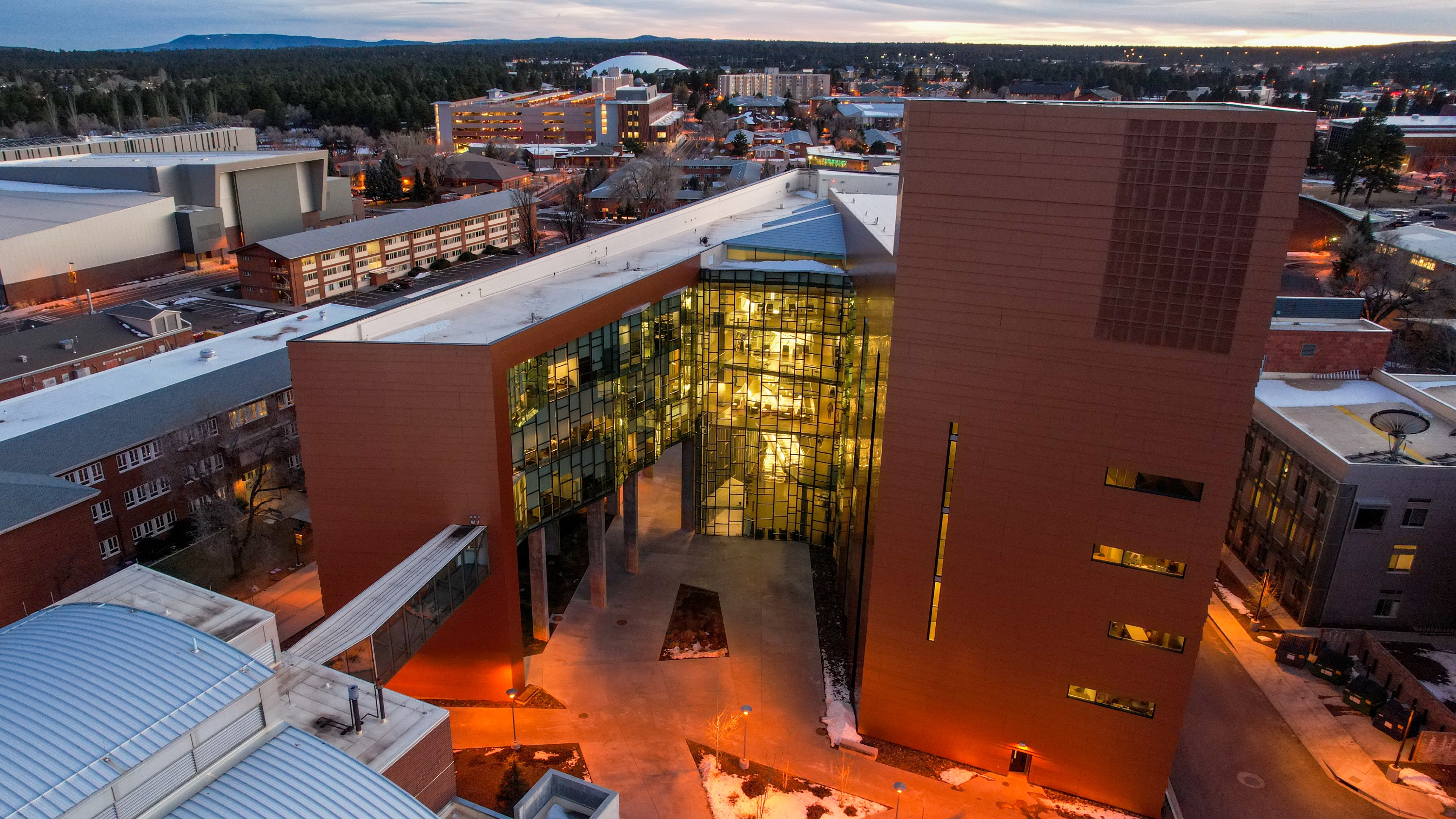 Aerial view of the N A U campus skyline at sunset, with a large, illuminated glass and steel building in the foreground.