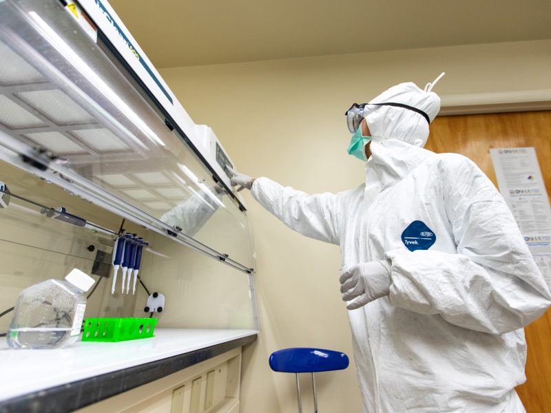 Person in protective medical hazmat suit working in a laboratory setting with scientific equipment and a monitor.