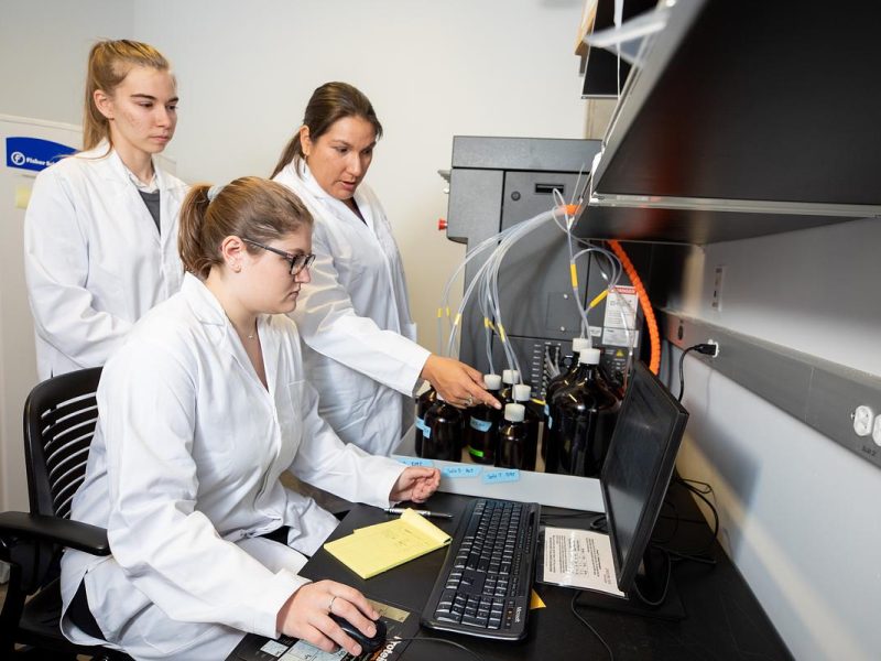 Three people in white lab coats standing in a laboratory, working with a computer and other equipment. One person is sitting at the computer, the others are standing nearby observing or assisting.