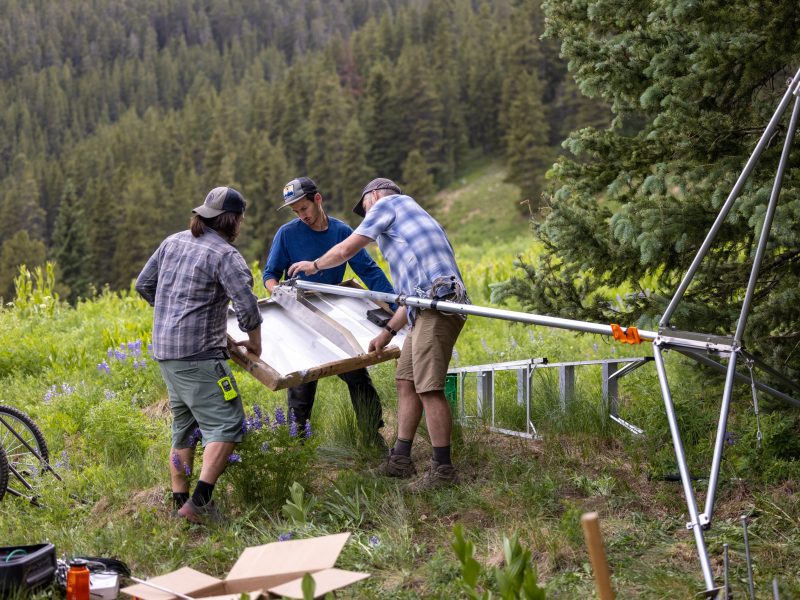Three people carrying a large object through a forested area, with mountains and trees in the background.