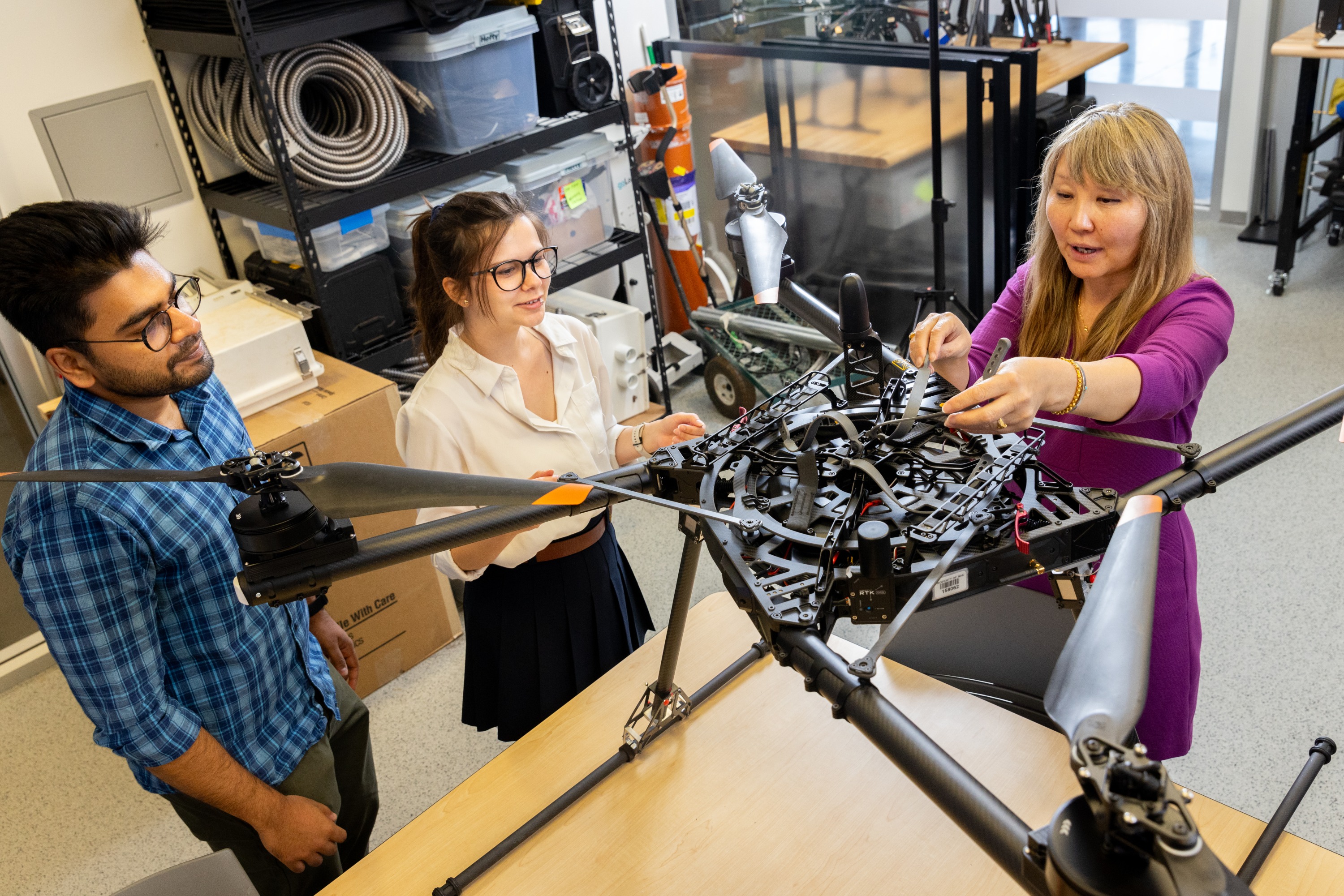 A group of three people working together on an industrial-looking device in a workshop or laboratory setting. The device appears to be a complex mechanical frame or piece of equipment. The individuals are shown examining and manipulating the device, suggesting they are engaged in some kind of collaborative work or project.
