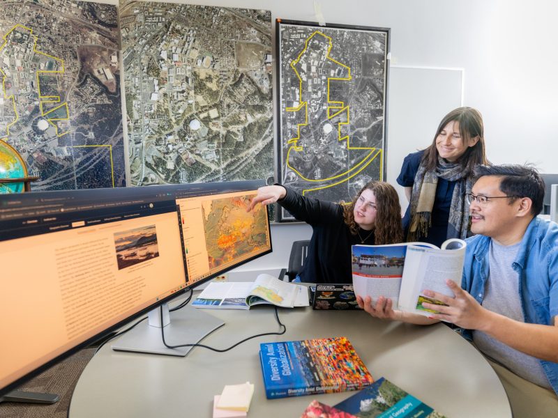 Three people in a professional office setting examining satellite imagery maps displayed on computer monitors. Large aerial maps with yellow highlighting are mounted on the wall behind them. The desk contains satellite imagery documentation, reference materials, and a desktop computer setup. The monitors show thermal or satellite data visualizations in orange and red tones. A globe is visible on the left side of the desk.